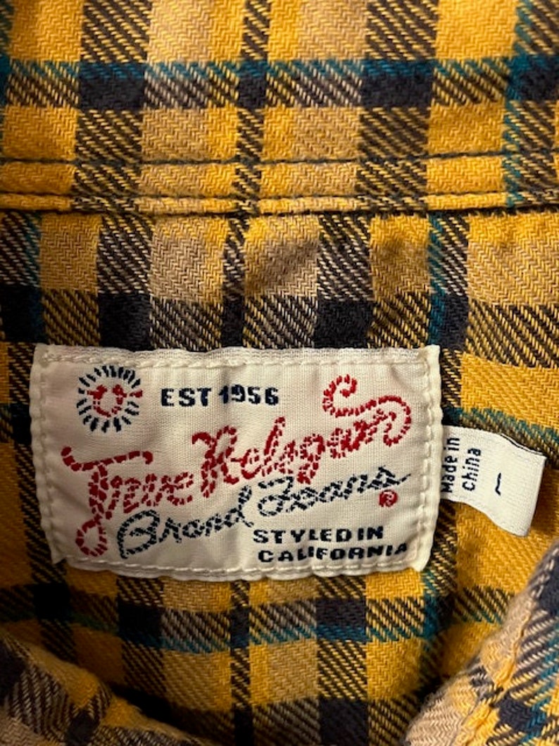 Puede incluir: Una camisa de cuadros amarilla, negra y azul con una etiqueta blanca que dice "True Religion Brand Jeans" y "EST 1956" en letras rojas. La etiqueta tambi&eacute;n dice "Styled in California" y "Made in China".