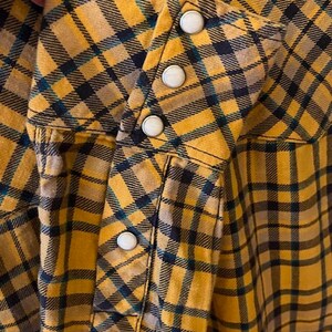 Puede incluir: Una camisa de cuadros amarillos y negros con cierre de botones. La camisa tiene manga larga y un bolsillo en el lado izquierdo.