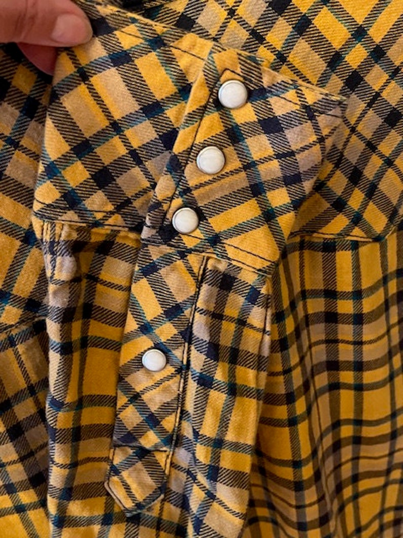 Puede incluir: Una camisa de cuadros amarillos y negros con cierre de botones. La camisa tiene manga larga y un bolsillo en el lado izquierdo.