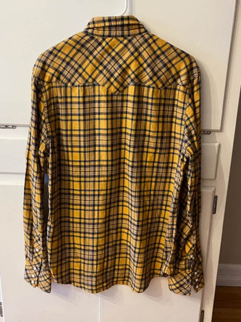 Puede incluir: Una camisa de manga larga a cuadros amarillos y negros con cuello puntiagudo y un bolsillo en el pecho.