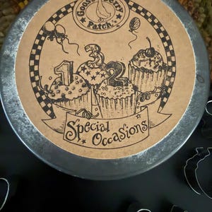 May include: A round metal tin with a tan paper label featuring a black ink illustration of three cupcakes topped with the numbers 1, 3, and 2. The label includes the text Gooseberry Patch and Special Occasions, surrounded by balloons.