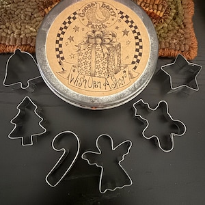 May include: A round metal tin with a tan label featuring a gift box illustration and the text Gooseberry Patch Wish Upon A Star. Surrounding the tin are six silver metal cookie cutters in the shapes of a bell, star, tree, candy cane, angel, and gingerbread person.