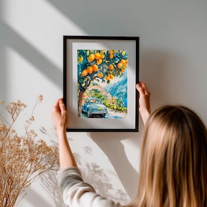 May include: A framed watercolor painting depicts a scenic Italian landscape. A light blue vintage car drives on a road lined with buildings, under a tree laden with oranges. The artwork is framed in a black frame and is being held up against a white wall.