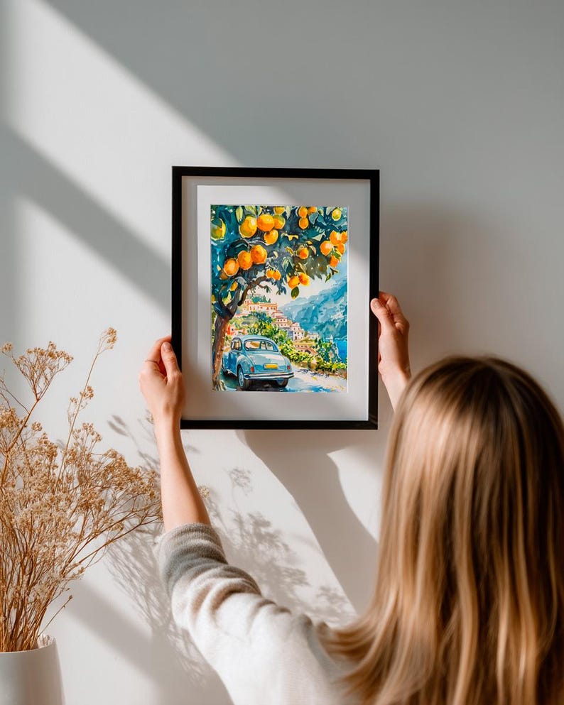May include: A framed watercolor painting depicts a scenic Italian landscape. A light blue vintage car drives on a road lined with buildings, under a tree laden with oranges. The artwork is framed in a black frame and is being held up against a white wall.