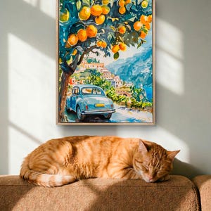 May include: A framed watercolor painting depicts a coastal Italian scene with a blue car parked under an orange tree. A sleeping orange tabby cat rests on a brown sofa below the artwork, bathed in sunlight.