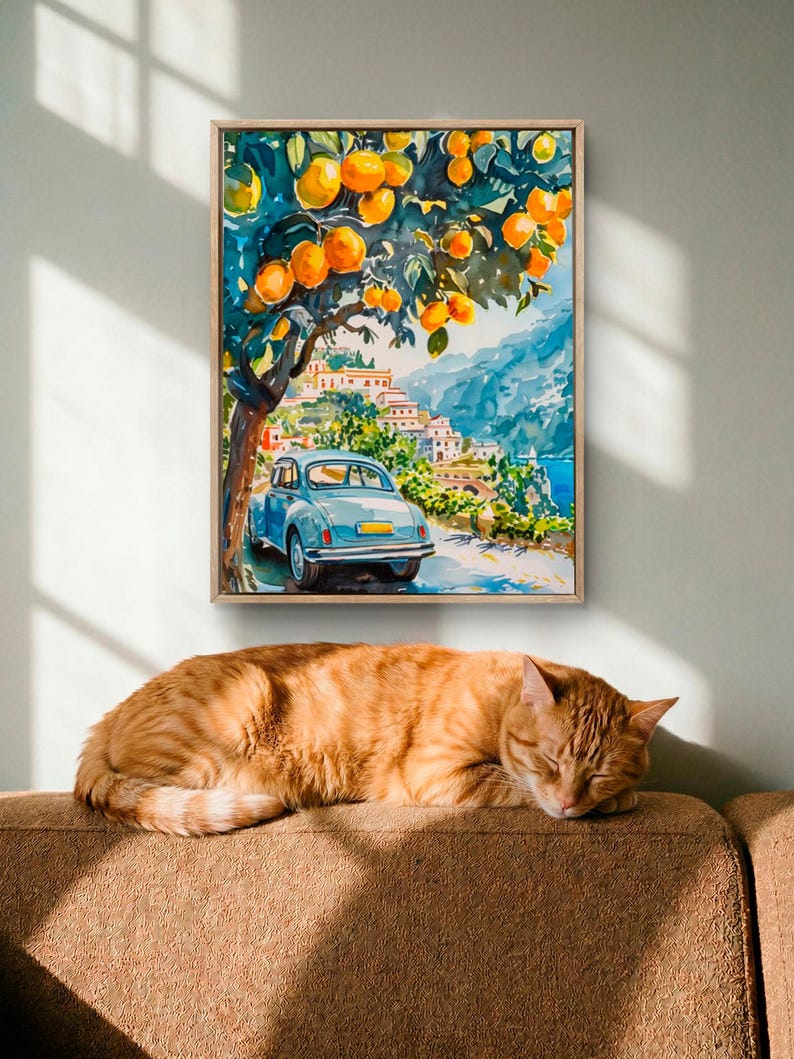 May include: A framed watercolor painting depicts a coastal Italian scene with a blue car parked under an orange tree. A sleeping orange tabby cat rests on a brown sofa below the artwork, bathed in sunlight.