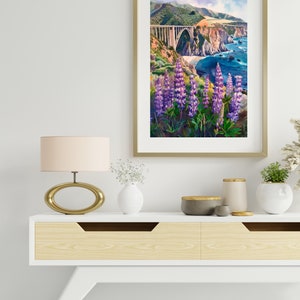 May include: A framed watercolor painting of a bridge over a body of water with purple flowers in the foreground. The bridge is a concrete arch bridge with a green hillside behind it. The water is blue and there are white waves crashing on the shore.