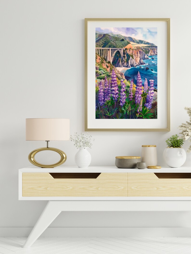 May include: A framed watercolor painting of a bridge over a body of water with purple flowers in the foreground. The bridge is a concrete arch bridge with a green hillside behind it. The water is blue and there are white waves crashing on the shore.