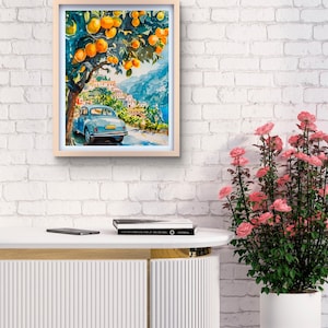 May include: Watercolor painting in a light wood frame. The artwork depicts a blue vintage car on a road, under an orange tree, with a coastal town in the background. The painting has a bright, sunny aesthetic.