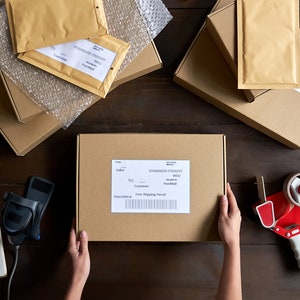 May include: A flat lay image of a variety of brown cardboard boxes and bubble wrap envelopes, some with labels and barcodes. A person's hands are holding a brown box with a shipping label that reads "Standard Postage". A red and white tape dispenser is on the table.