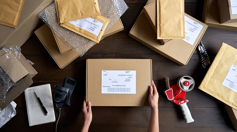 May include: A flat lay image of a variety of brown cardboard boxes and bubble wrap envelopes, some with labels and barcodes. A person's hands are holding a brown box with a shipping label that reads "Standard Postage". A red and white tape dispenser is on the table.