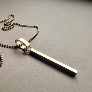 May include: A gold-colored bar pendant necklace with a ball chain. The pendant is a rectangular bar suspended from a small ring. The chain and pendant have a metallic finish, set against a dark background.