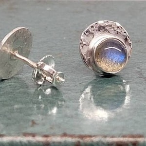 Puede incluir: Un par de pendientes de plata con una piedra preciosa redonda azul iridiscente engastada en un engaste de plata texturizado. Los pendientes tienen un poste y una mariposa en la parte posterior.