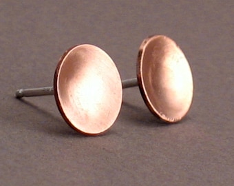 Puede incluir: Un par de pendientes de botón de cobre con una forma redondeada ligeramente abovedada. Los pendientes tienen una espiga de plata.