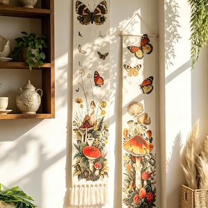 May include: Two vertical fabric wall hangings featuring botanical illustrations of mushrooms and butterflies. The off-white banners are suspended from wooden rods with white string and finished with cream-colored fringe at the bottom edges.