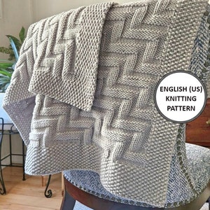 May include: A gray knitted blanket with a woven pattern. The blanket is folded and draped over a chair. The text "ENGLISH (US) KNITTING PATTERN" is visible in the lower right corner of the image.