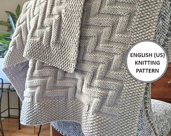 Op de afbeelding: Een grijs gebreid deken met een geweven patroon. Het deken is gevouwen en over een stoel gelegd. De tekst "ENGLISH (US) KNITTING PATTERN" is zichtbaar in de rechterbenedenhoek van de afbeelding.