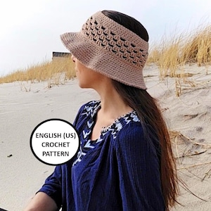 May include: A crocheted visor in a light tan color with a decorative openwork pattern. The visor has a brim and a band that fits around the head. The image also includes the text "ENGLISH (US) CROCHET PATTERN".