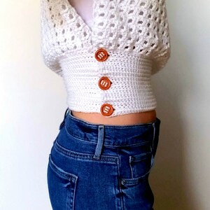 May include: A white crocheted crop top with a V-neck and brown buttons. The top is worn over blue jeans.