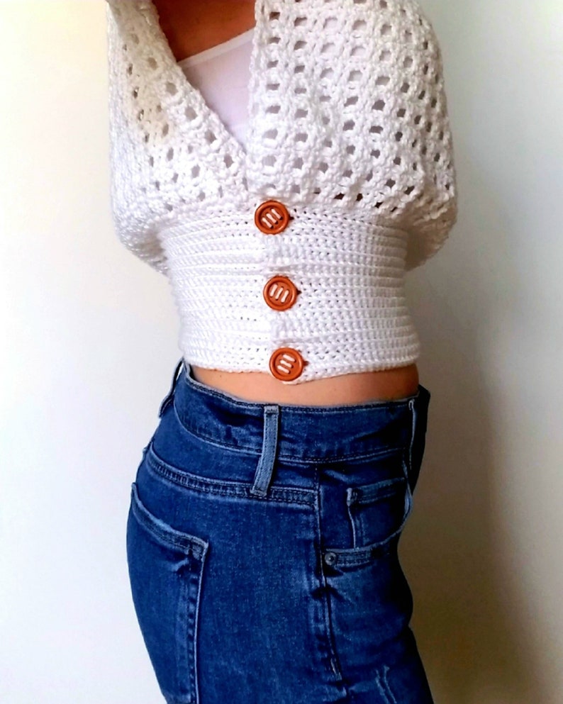 May include: A white crocheted crop top with a V-neck and brown buttons. The top is worn over blue jeans.