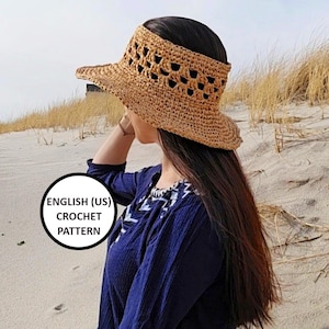 Può includere: Un cappello da sole all'uncinetto con un'ampia tesa e un design a uncinetto. Il cappello è fatto di paglia naturale e ha un colore marrone chiaro. Il cappello viene indossato da una persona con lunghi capelli scuri. Il cappello viene indossato su una spiaggia con sabbia ed erba sullo sfondo. Il testo "ENGLISH (US) CROCHET PATTERN" è visibile nell'immagine.