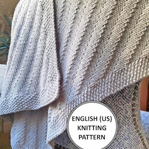 以下が含まれることがあります： グレーの編み地の毛布で、シェブロン模様が施されています。毛布は半分に折り畳まれ、椅子にかけられています。毛布の円の中に「ENGLISH (US) KNITTING PATTERN」というテキストが表示されています。