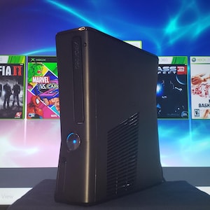 May include: Black Xbox 360 console with a blue power button, positioned in front of a screen displaying game cases. The game cases feature titles like "Mafia II" and "Marvel vs. Capcom". The screen has a blue and white gradient.