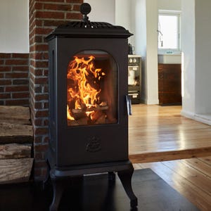May include: A black cast iron stove with a glass door showing a burning fire. The stove has decorative details and stands on four curved legs. A brick wall and wooden floor are in the background.