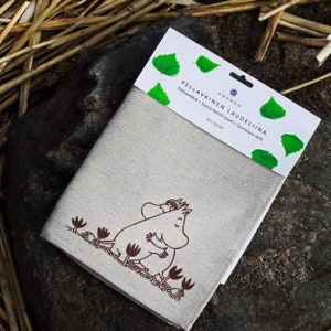 May include: A beige linen sauna bench towel with a brown illustration of two Moomins hugging. The towel is labeled "Pellavainen Laudeliina" and "Sitthandduk - Sauna bench towel - Saunalava rätik" in Finnish. The towel measures 50 x 150 cm.