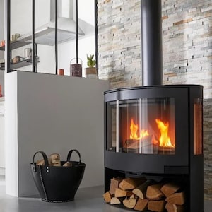 May include: A contemporary black fireplace with a glass front, displaying burning flames and stacked firewood. A black leather basket filled with logs is positioned nearby. The fireplace is set against a stone wall, creating a warm atmosphere.