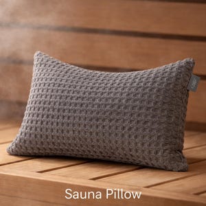Può includere: Un cuscino da sauna grigio con una trama a nido d'ape. Il cuscino rettangolare è esposto su una superficie di legno. Le parole "Sauna Pillow" sono stampate in basso nell'immagine.