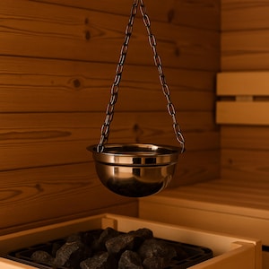 May include: A stainless steel sauna bucket suspended by a chain, positioned above a tray of dark rocks. The sauna setting features wooden walls and benches, creating a warm, inviting atmosphere for relaxation and wellness.