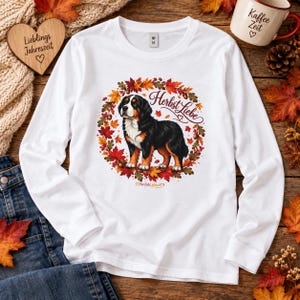 Könnte beinhalten: Ein weißes Langarmshirt mit einer Grafik eines Berner Sennenhundes, der von einem kreisförmigen Kranz aus bunten Herbstblättern und Eicheln umgeben ist. Der Schriftzug "Herbst Liebe" ist in Schreibschrift über dem Hund abgebildet.