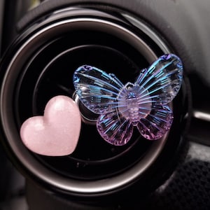 May include: A car air vent clip featuring a translucent butterfly with iridescent blue and purple hues, and a pink heart-shaped charm. The butterfly is positioned near the vent, adding a decorative touch to the car's interior.