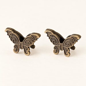 May include: A pair of butterfly-shaped metal objects with an antique brass finish. Each piece features a raised, dark-toned scrollwork pattern on the wings, creating a textured appearance. The butterfly design is symmetrical with curved edges.