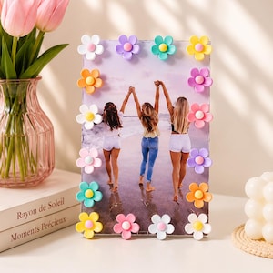 May include: A rectangular picture frame decorated with colourful plastic flower embellishments in shades of white, yellow, orange, pink, purple, and teal. The frame holds a photograph of three people walking on a beach, viewed from behind.