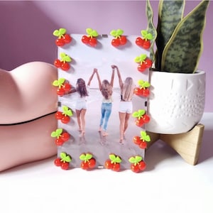 May include: A clear acrylic photo frame decorated with red cherry accents and green leaves. The frame holds a photo of three people on a beach. A white ceramic pot with a plant sits on a wooden stand.