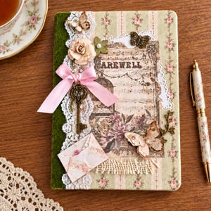 Könnte beinhalten: Ein rechteckiges Notizbuch mit floralem Streifenmuster, verziert mit einer rosa Satinschleife, einem Metallskelettschlüssel, Spitzenbesatz, Papierblumen, Schmetterlingsapplikationen und einem kleinen Umschlag. Der Einband trägt den Text FAREWELL.