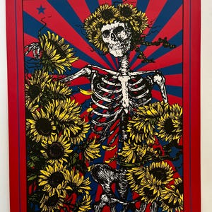 May include: A colourful print depicting a skeleton wearing a sunflower crown and surrounded by sunflowers. The background has red and blue rays with a star. The artwork is framed in red.