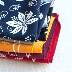 May include: A stack of five fabric squares with different patterns. The top square is navy blue with a white floral design. The second square is gray with a white floral design. The third square is orange with a white floral design. The fourth square is burgundy with a white floral design. The bottom square is red with a white floral design.
