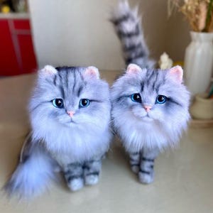 May include: Two plush cat figurines with long, fluffy grey and white striped fur, bright blue eyes, and small pink noses. The cats have pointed ears, detailed facial features, and long, bushy tails, standing in a realistic, upright posture.