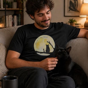 May include: Black t-shirt with a graphic of a cat silhouette on a branch in front of a yellow moon, with the text "Fly me to the Moon". The shirt is worn by a person sitting on a couch with a black cat.