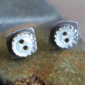 May include: A pair of silver earrings with a white button design. Each earring features a square-shaped setting with a white button in the center. The button has two holes and a scalloped edge.