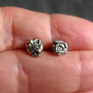 May include: A pair of small, silver-colored stud earrings. Each earring has a textured, irregular surface, resembling a small nugget or a cluster of tiny shapes. The earrings are shown against a blurred background.