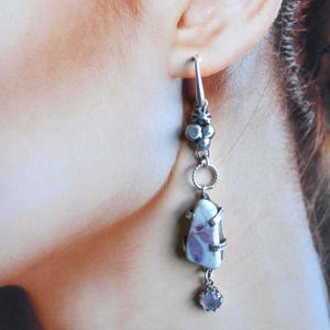 May include: A long drop earring featuring a silver hook, a cluster of small silver spheres, a twisted silver ring, and a central irregular white stone with purple inclusions held by silver prongs, finished with a small dangling round stone.