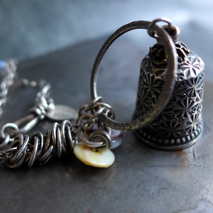 May include: A silver-toned thimble pendant featuring an intricate starburst pattern, attached to a textured metal ring. The piece includes a silver chain with coiled wire accents and small, iridescent, circular shell charms dangling from the ring.