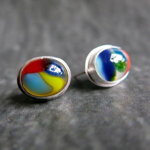 May include: A pair of oval stud earrings featuring smooth, multi-colored glass cabochons set in silver-toned metal frames. The glass displays swirling patterns of red, yellow, blue, and green, creating a vibrant, glossy surface on each earring.