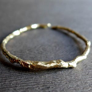 May include: A gold-colored ring with an open, circular band featuring a textured, uneven surface that mimics the appearance of a small, thin twig or branch. The metal has a reflective, metallic finish with visible ridges and bumps along the entire circumference of the band.