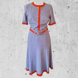 Könnte beinhalten: Ein graues Kleid mit rotem Besatz an Ausschnitt, Ärmeln und Saum. Das Kleid hat einen Knopfleisten vorne und einen Gürtel mit einer silbernen Schnalle.