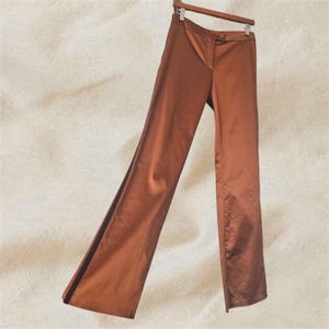 Può includere: Pantaloni di raso marroni appesi a una gruccia di legno. I pantaloni hanno una chiusura con bottoni e un design a gamba dritta. Il tessuto ha una leggera lucentezza e i pantaloni sembrano di peso medio. I pantaloni sono di colore uniforme e adatti a varie occasioni.